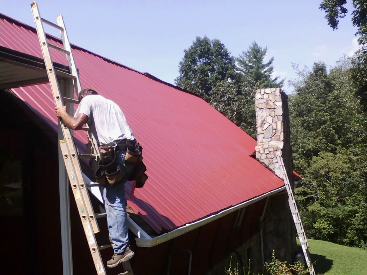Roofer on a ladder beside a red metal roof and stone chimney — Soffit & Fascia Repair by Roofing Brothers in Franklin